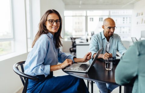 Mature female designer sitting around a table and smiling at the camera. Happy business woman having a meeting with her team in an office.